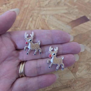 May include: Two embroidered patches of a reindeer with a red nose, white antlers, and black hooves. The reindeer are tan with white accents. The patches are on a person's hand.