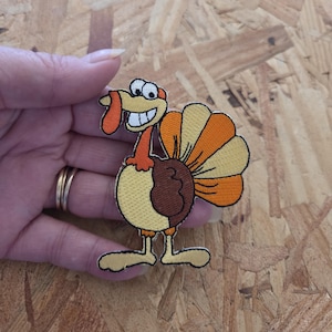 May include: A colorful embroidered turkey patch with a smiling face, orange wattle, and a fan of orange and yellow feathers. The turkey has a brown body and yellow legs. The patch is held in a hand.