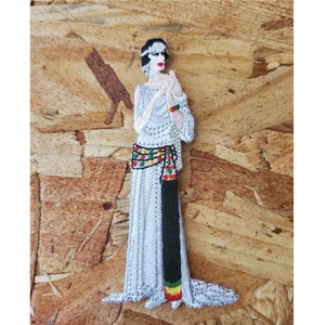 May include: An embroidered iron-on patch featuring a woman in a white dress with a colorful sash and a black and red striped sash. The woman has black hair and a white face with red lips.