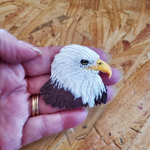 May include: Embroidered patch featuring a detailed bald eagle head. The eagle has white feathers, a brown neck, a yellow beak, and a focused gaze. The patch is likely for clothing or accessories.