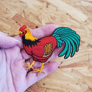 May include: Embroidered patch of a rooster with a red body, yellow head, black accents, and a teal tail. The rooster has yellow legs and a red comb. The patch is likely for clothing or accessories.