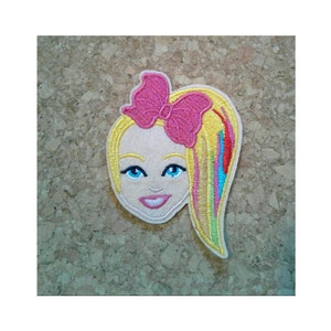 May include: Embroidered iron-on patch featuring a cartoon girl with blonde hair, a pink bow, and a colorful rainbow streak in her hair.