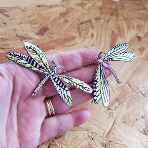 May include: Two dragonfly patches with embroidered details. The wings are a mix of black, white, pink, and green, with delicate patterns. The dragonflies are held in a hand, showcasing the intricate design and craftsmanship.