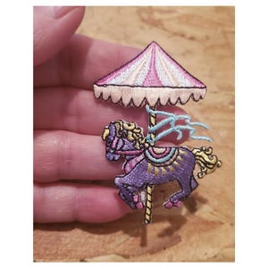 May include: An embroidered iron-on patch of a purple carousel horse with a pink and white striped canopy. The horse has a blue and yellow saddle and a gold mane and tail.