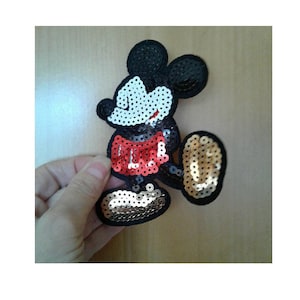 May include: A sequined Mickey Mouse patch. The patch features a black body, ears, and outline. The face is white, the shorts are red, and the feet are gold. The sequins give the patch a shiny appearance.