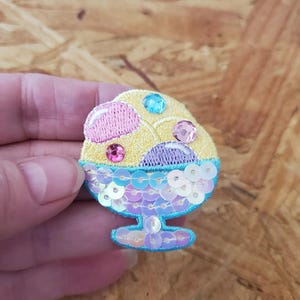 May include: Embroidered ice cream sundae patch with yellow, pink, and blue colors. The ice cream has pink and yellow scoops with blue and pink gems. The sundae dish is blue with iridescent sequins.