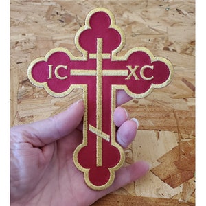 May include: Red embroidered cross patch with gold trim and the letters "IC XC" in gold. The cross is on a wooden background.