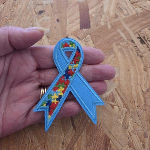 May include: An embroidered awareness ribbon patch featuring a light blue border and a central section filled with a colorful puzzle piece pattern in red, yellow, green, and blue. The ribbon has a classic looped shape with two pointed tails at the bottom.