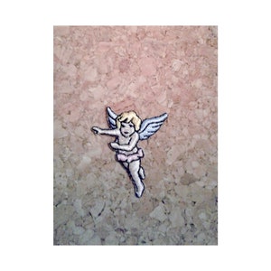May include: A small embroidered patch of a cherub with blonde hair, pink wings, and a pink skirt. The cherub is flying with its arms outstretched.