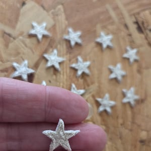 May include: A collection of small, embroidered star patches in a light silver color. The stars are arranged on a wooden surface, with one held between fingers. These patches are ideal for crafts and embellishments.