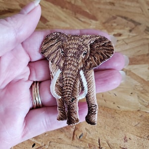 May include: Embroidered elephant patch in shades of brown and tan, featuring detailed stitching. The patch has white tusks and is held in a hand. This patch is suitable for clothing, bags, or other fabric items.