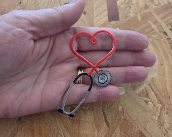 May include: An embroidered patch featuring a stethoscope with its tubing shaped into a heart outline. The heart is bright red, while the stethoscope chest piece and earpieces are rendered in shades of grey and black with detailed stitching.