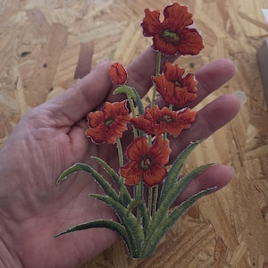 May include: Embroidered patch featuring a cluster of red poppy flowers with green centers and stems. The patch has detailed stitching and green leaves. The flowers are in various stages of bloom, with some buds visible.