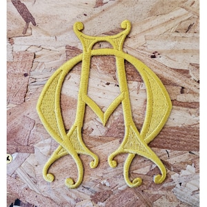 May include: A gold embroidered patch with a stylized letter 'M' design. The patch is on a wood background.