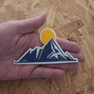 May include: Embroidered patch featuring a mountain range with a yellow sun. The patch has a navy blue mountain design with white and green accents, and a white border. The patch is held in a hand.