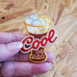 May include: Embroidered patch in the shape of a beverage with the word "Cool" in red and silver. The patch features brown, yellow, and white accents, with sequins adding sparkle. The design resembles a refreshing drink.