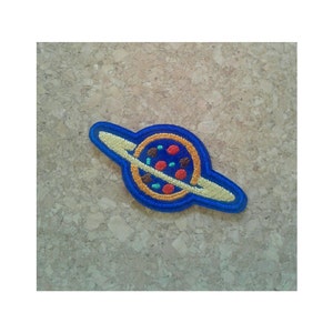 May include: A blue and yellow embroidered patch of a pizza planet with a ring around it.