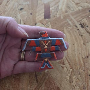 May include: Embroidered patch featuring a stylized bird design. The patch has a brown, orange, and blue color scheme. The bird's wings are spread, and it has a geometric pattern on its body. The patch is likely intended for application on clothing or accessories.