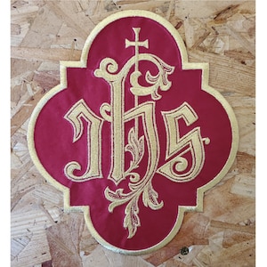 May include: A red embroidered patch with gold trim and the letters IHS, topped with a cross. The patch has a quatrefoil shape. The letters are in an ornate, stylized font with decorative flourishes.