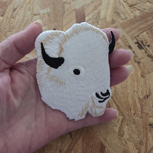 May include: Embroidered patch featuring a white bison head with black horns and eye. The bison's fur is detailed with beige stitching. This patch is likely designed for application on clothing or accessories.