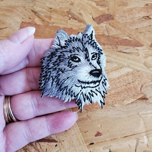 May include: Embroidered wolf patch in shades of gray and black. The wolf's face is intricately detailed, with a focus on the eyes, nose, and fur texture. The patch is held in a hand, showcasing the size and detail of the artwork.