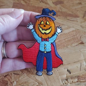 May include: Embroidered patch featuring a smiling pumpkin head wearing a blue witch hat and cape. The pumpkin is dressed in a blue shirt, pants, and a red bow tie. The patch is designed for Halloween.