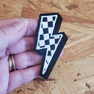 May include: Black and white lightning bolt patch with a checkered pattern. The patch has a white border and is held in a hand. The background is a light brown wooden surface.
