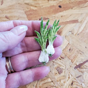 May include: Two embroidered onion-like plant appliques. The appliques feature white bulbous bottoms and green and yellow tops. The appliques are held in a hand with multiple rings. The background is a light brown wood.