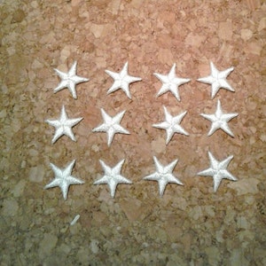 May include: Twelve white embroidered star-shaped patches on a corkboard background.