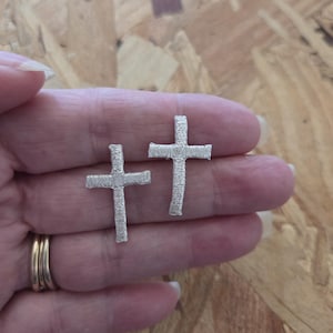 May include: A pair of silver-colored cross earrings. The earrings are small, with a textured surface, and are held in a hand. The crosses are a religious symbol, and the earrings are likely for everyday wear.