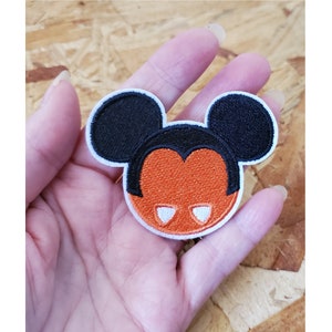 May include: Orange and black embroidered iron-on patch of a Mickey Mouse head with white eyes and fangs.
