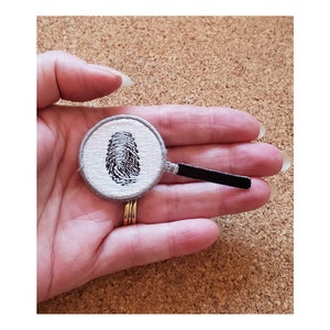 May include: A white embroidered patch with a black fingerprint inside a magnifying glass. The magnifying glass has a black handle.
