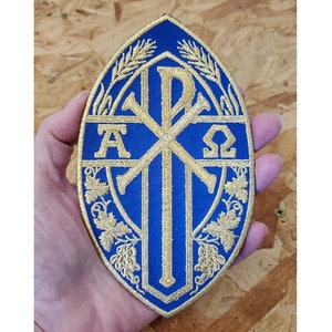 May include: Blue and gold embroidered patch with a cross, the letters Alpha and Omega, and wheat and grape vines. The patch is oval shaped.