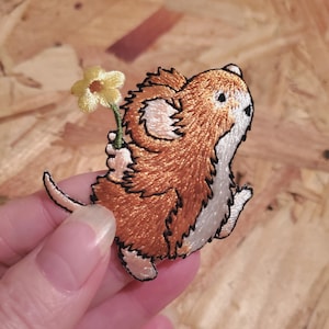 May include: Embroidered iron-on patch of a brown mouse holding a yellow flower. The mouse is facing right and has a white belly. The patch is perfect for adding a whimsical touch to clothing, bags, or other fabric items.