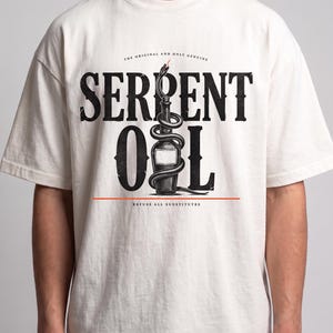 Peut inclure: Un t-shirt à manches courtes blanc cassé avec un motif noir représentant une bouteille entourée d'un serpent. Le texte indique "THE ORIGINAL AND ONLY GENUINE", "SERPENT OIL" et "REFUSE ALL SUBSTITUTES" dans une police de style vintage.