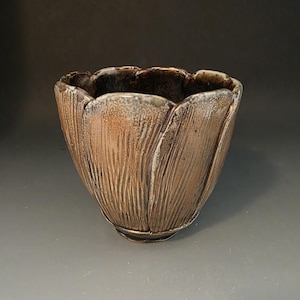 May include: A ceramic bowl with a scalloped, petal-like rim and a textured, vertical-ribbed exterior. The surface features a metallic bronze and copper glaze with dark brown accents, while the interior is a deep, dark brown with a smooth finish.