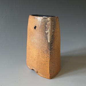 May include: A tall, hand-built ceramic vase with a textured, mottled orange-brown surface. The vessel features a flat, angular top edge, a dark interior rim, and a single small circular perforation near the upper left side of the exterior wall.