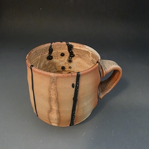 May include: A handmade ceramic mug with a warm tan and beige speckled finish, featuring vertical black drip lines that extend from the rim down the exterior and into the interior. The mug has a thick, curved handle and a rounded, organic shape.