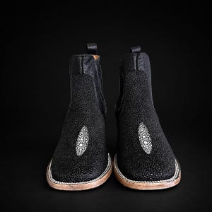 Könnte beinhalten: Ein Paar schwarze Stiefel mit einer texturierten Rochenleder-Optik und einem markanten weißen ovalen Einsatz auf dem Vorderblatt. Die Stiefel haben eine quadratische Zehenpartie, eine Zuglasche an der Ferse und einen hellen Rahmen mit sichtbaren Nähten.