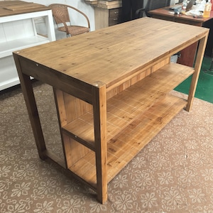 May include: A rectangular wooden kitchen island featuring a smooth top surface and two open shelves underneath for storage. The piece is constructed from medium-toned brown wood with visible grain patterns and four sturdy vertical legs at each corner.
