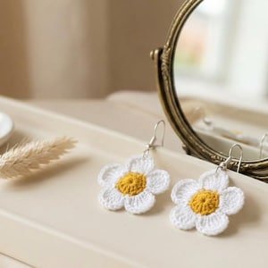 May include: A pair of handmade crochet flower earrings featuring white petals and a yellow center, attached to silver-toned metal hooks. The earrings are displayed alongside a small round mirror and a dried wheat stalk on a neutral-colored tray.