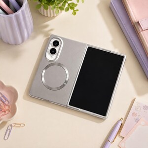 May include: A silver foldable smartphone case featuring a circular magnetic ring on the back panel and a dual-camera module with a white border. The case has a smooth matte finish and is designed to protect a device with a folding screen mechanism.