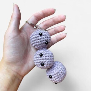 May include: A lavender crochet keychain featuring three spherical, interconnected shapes. Each sphere is crafted from yarn and includes two small, round, black plastic eyes. The keychain is attached to a standard silver-colored metal split ring.