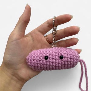 May include: A light pink, cylindrical crochet keychain featuring two small, round, black plastic eyes on the front. The plush, textured yarn object is attached to a silver-toned metal split ring and chain, with a short pink yarn loop hanging down.