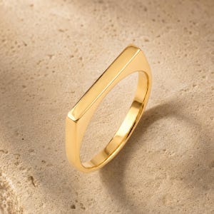 May include: A polished gold ring featuring a flat, rectangular bar top section. The band is smooth and continuous, showcasing a reflective metallic finish. The design is simple and clean, highlighting the solid gold construction and the geometric shape.