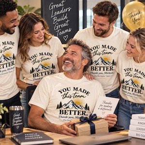 Può includere: T-shirt color bianco sporco con una grafica raffigurante una catena montuosa, un sole e pini, con il testo "YOU MADE this team BETTER. Thank you, Michael. The Northstar Team" stampato in inchiostro nero e oro sul petto.