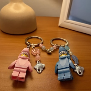 Pode incluir: Dois porta-chaves com minifiguras de plástico vestidas com fatos de tubarão, um em rosa claro e outro em azul claro. Cada porta-chaves inclui um anel de metal, um pendente de raia prateado e um fio de contas decorativas em tons pastel.