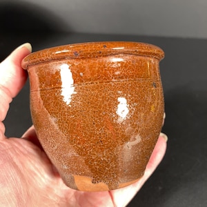 May include: A small, antique-style ceramic pot with a rich, brown glaze and a cracked, textured surface. The pot has a rounded shape with a slightly flared rim and horizontal bands. The pot is held in a hand.