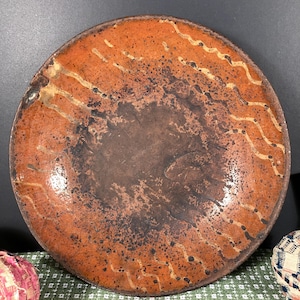 May include: A round, rustic ceramic plate with a mottled orange and brown glaze. The plate features a dark center and a lighter orange rim with wavy, cream-colored accents. The overall design suggests a handmade, artisanal aesthetic.