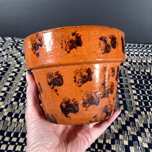 May include: An orange ceramic flower pot with dark brown splotches. The pot has a wide rim and a slightly tapered shape. The pot is held by a hand, showcasing its size and texture. The background is a blue and white patterned fabric.